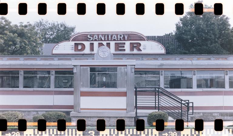 A diner with a sign that reads "Sanitary Diner" is visible from the street.