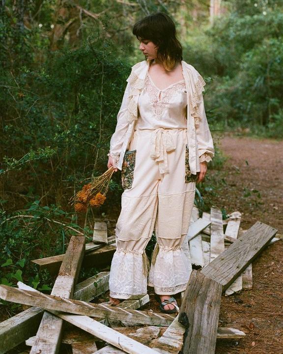 A woman in a cream-colored outfit with lace details stands on a pile of wooden planks in a forest, holding a bouquet of dried flowers.
