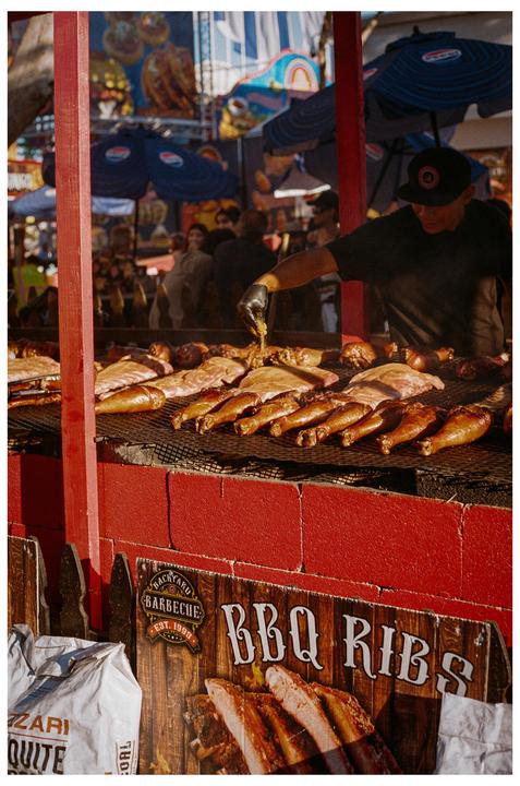 A man wearing a black cap and glove is basting meat on a large grill at a food stall.