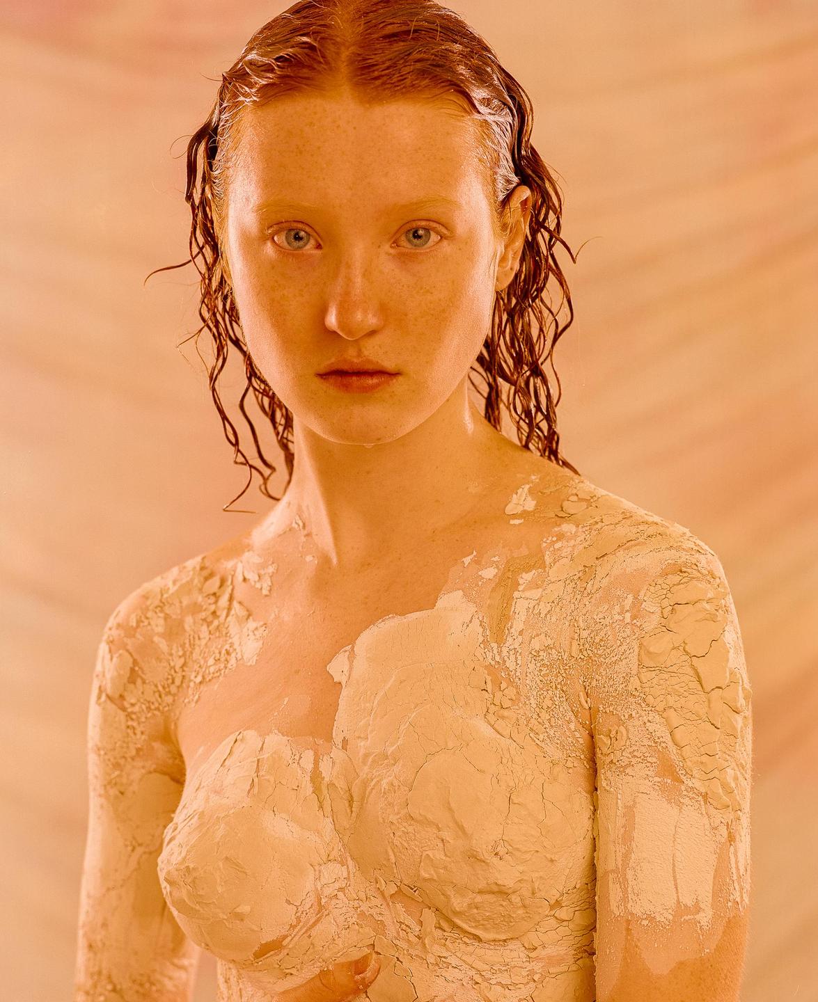A woman with wet, curly hair and clay on her body looks directly at the viewer.