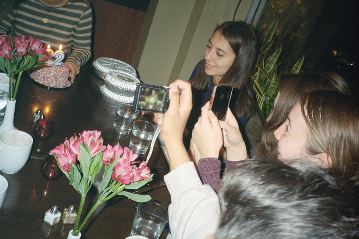 A group of women gather around a table with a birthday cake and flowers, with some of them holding up phones to take pictures.
