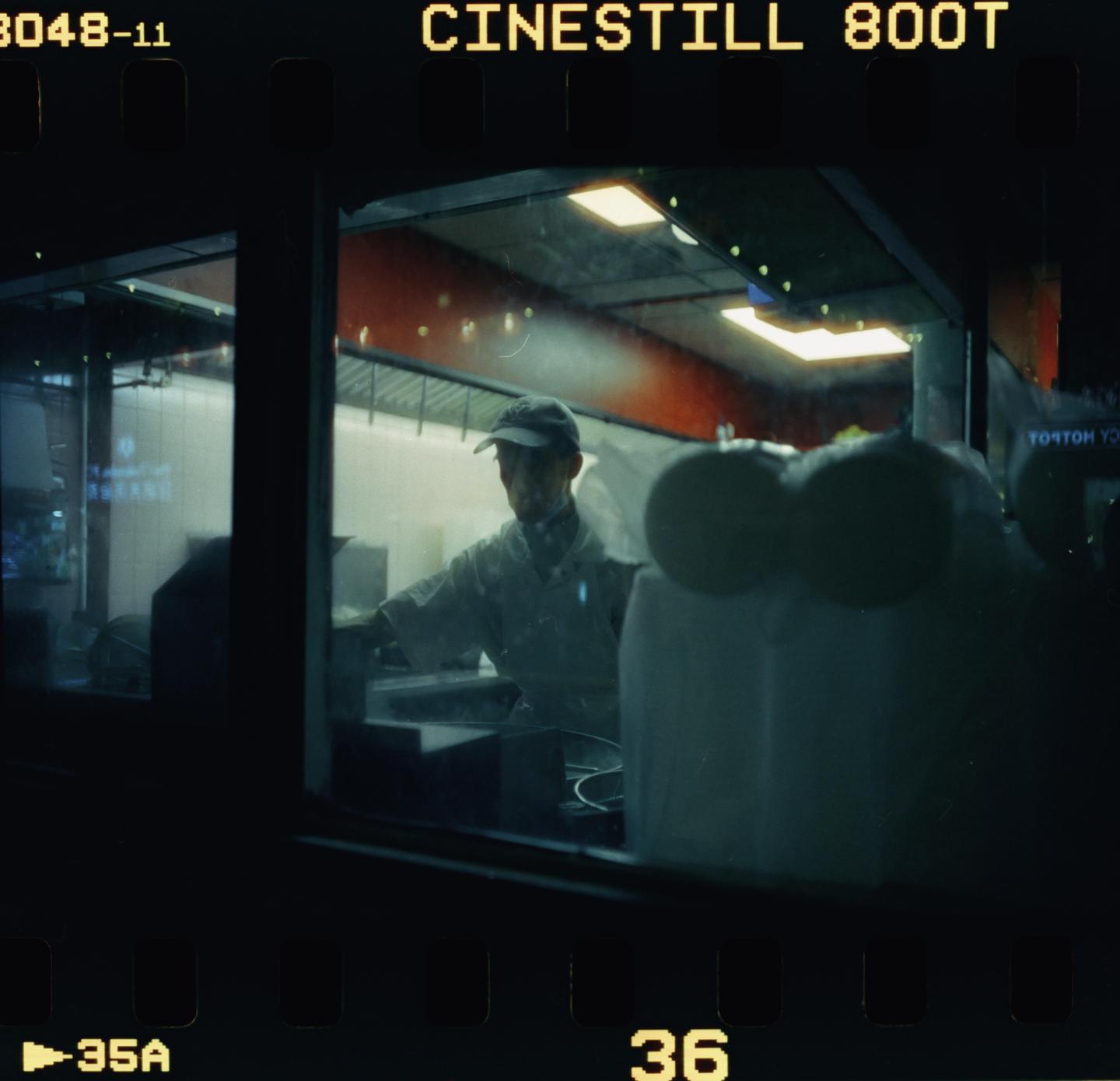 A man in a cap and white shirt works in a brightly lit kitchen at night, viewed through a window.