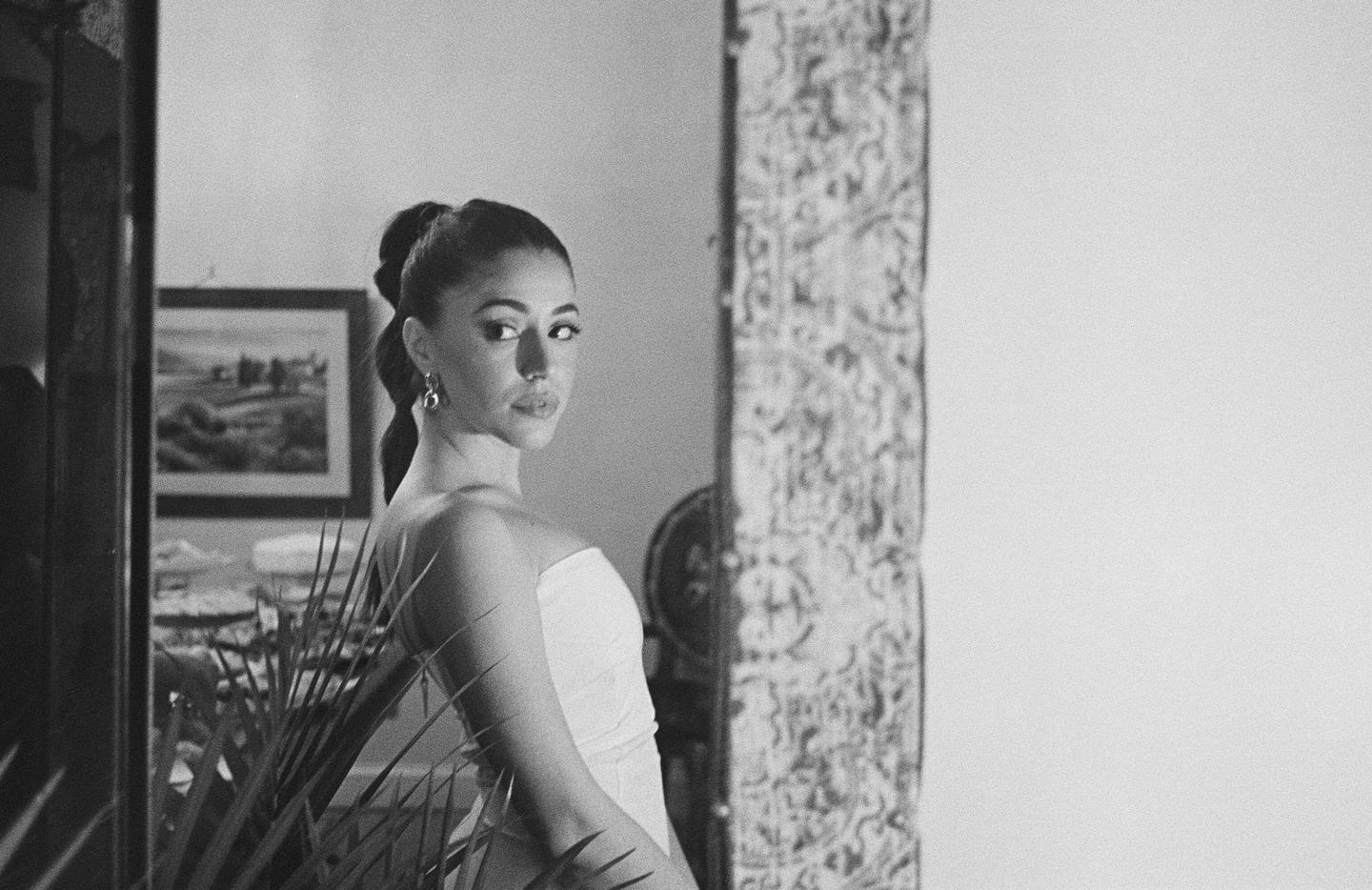 A woman with a ponytail looks over her shoulder at the viewer, with a framed picture and a plant in the background.
