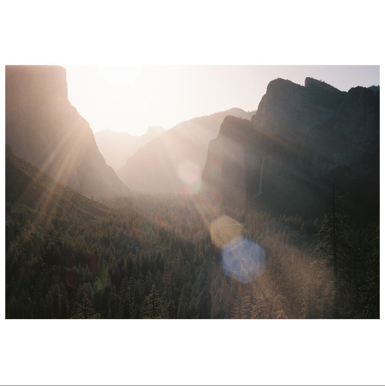 Sunlight streams over a valley filled with trees and surrounded by mountains.