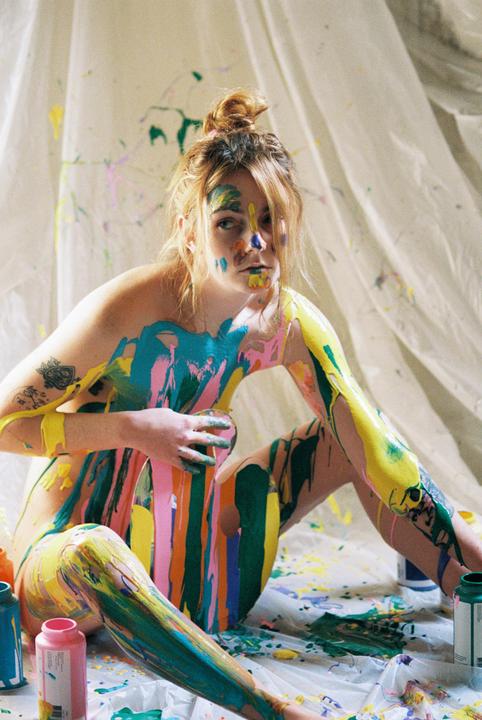 A woman sits covered in colorful paint with paint splatters on the white sheet behind her.