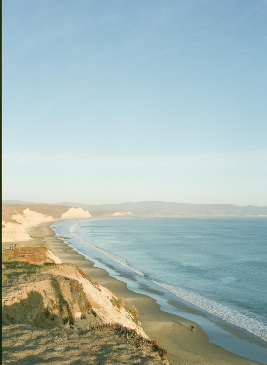 A long stretch of Drakes Beach is visible from a high vantage point, with waves breaking on the shore and cliffs rising on the left.