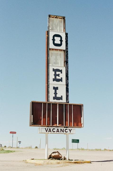 A tall, weathered motel sign with the word "MOTEL" vertically displayed and a "VACANCY" sign below stands against a clear blue sky.