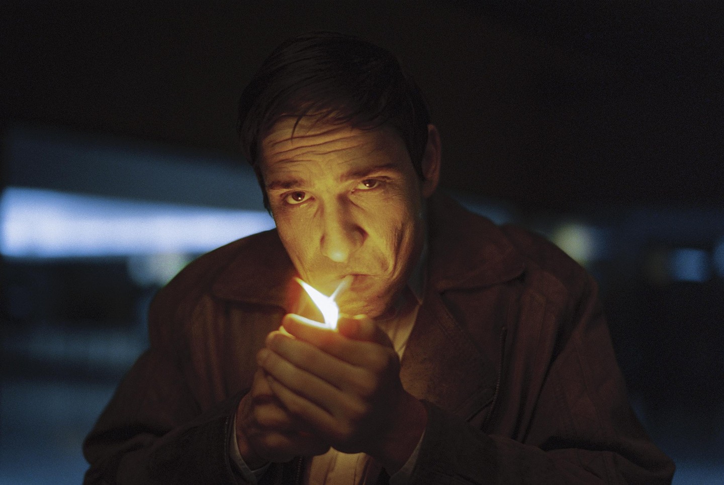 A man lights a cigarette in the dark, illuminated by the flame of a lighter.