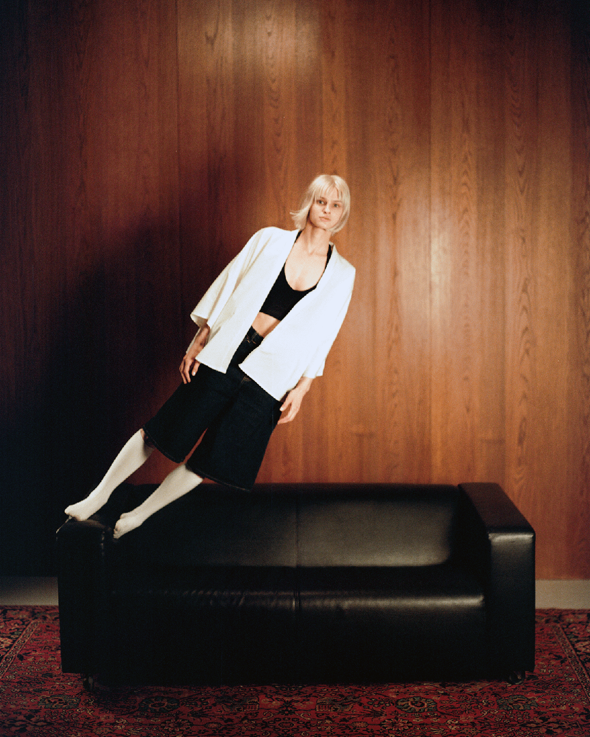 A woman with blonde hair is leaning against a wooden wall with her feet on a black sofa.