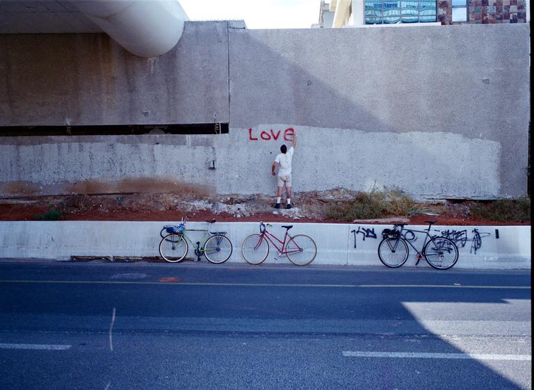 A man paints the word "Love" in red on a concrete wall next to a road with three bicycles parked along the curb.