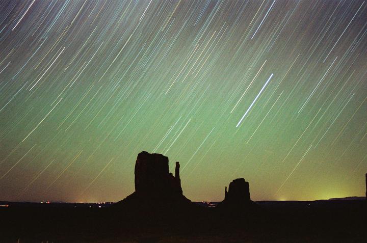 Two large rock formations are silhouetted against a night sky filled with star trails and a green glow.