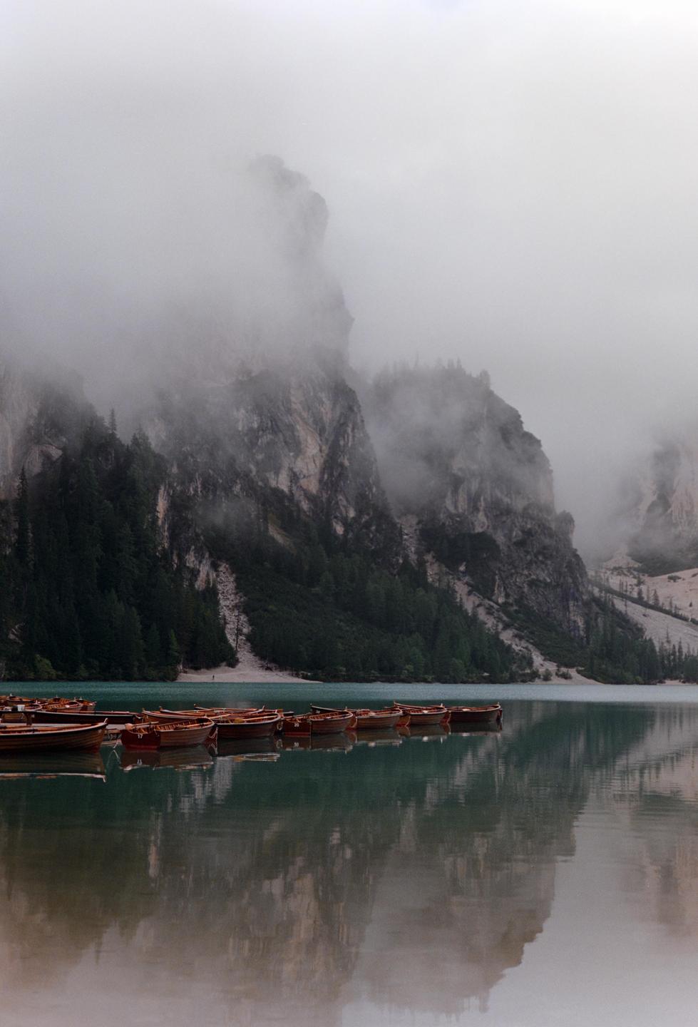 Wooden rowboats float on a lake surrounded by mountains shrouded in fog.
