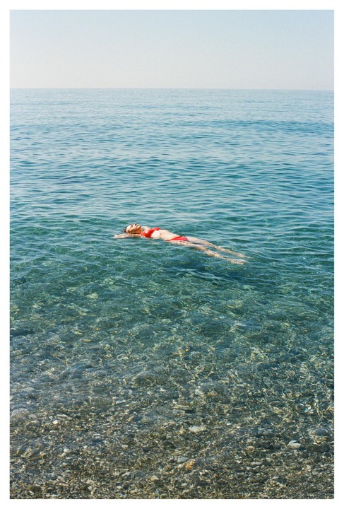 A woman in a red bikini floats on her back in clear blue water with a rocky bottom.