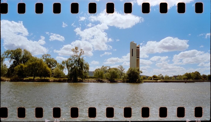 A body of water with trees and a tall, modern building on the far bank under a blue sky with white clouds.
