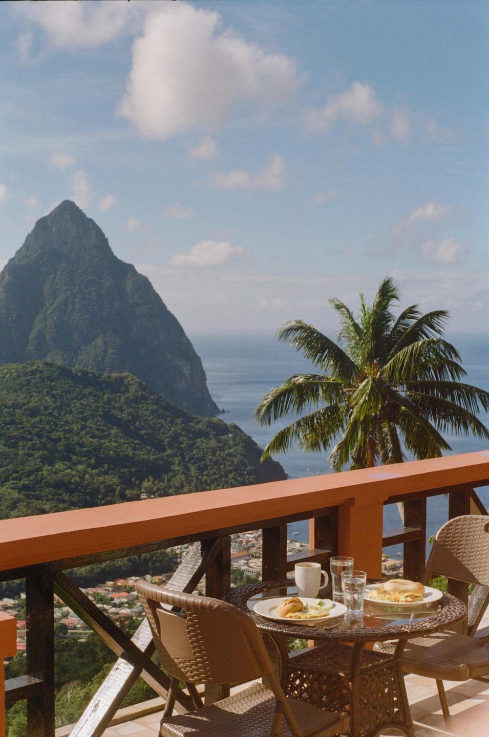 A breakfast table with food and drinks is set on a balcony overlooking a tropical bay with a large mountain and palm tree.