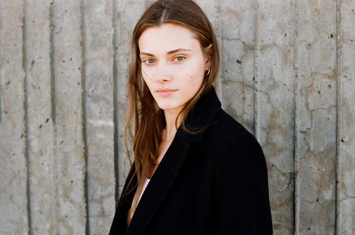 A woman with long hair and a black coat stands in front of a concrete wall.