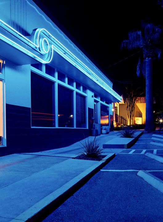 A building with blue neon lights on its facade stands next to a palm tree and a parking lot at night.