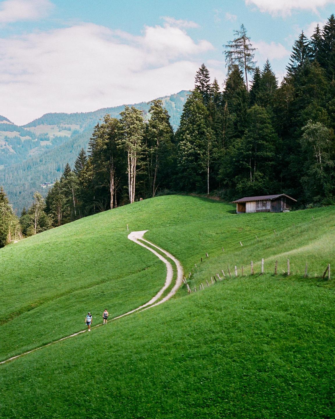 Two people walk on a winding dirt path through a green hillside with a small wooden hut and a forest in the background.