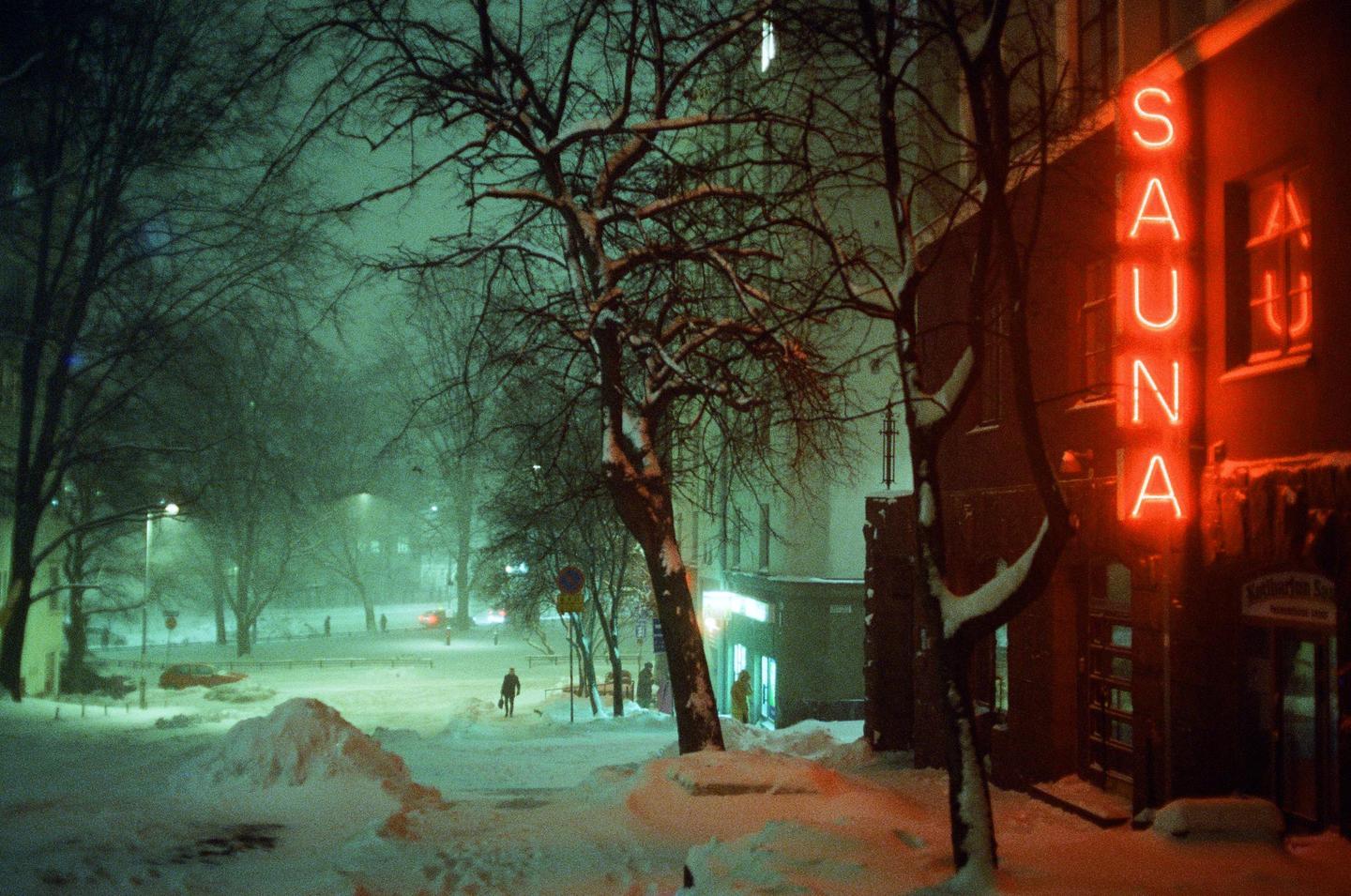 A neon sign for a sauna glows red on a snowy street at night, with bare trees and a person walking in the distance.