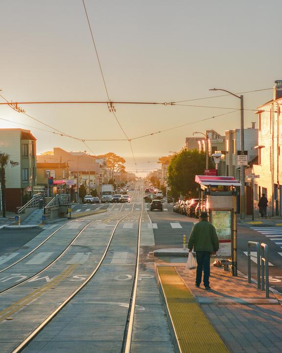 A man stands on a tram stop platform with a street and buildings stretching into the distance under a golden sky.