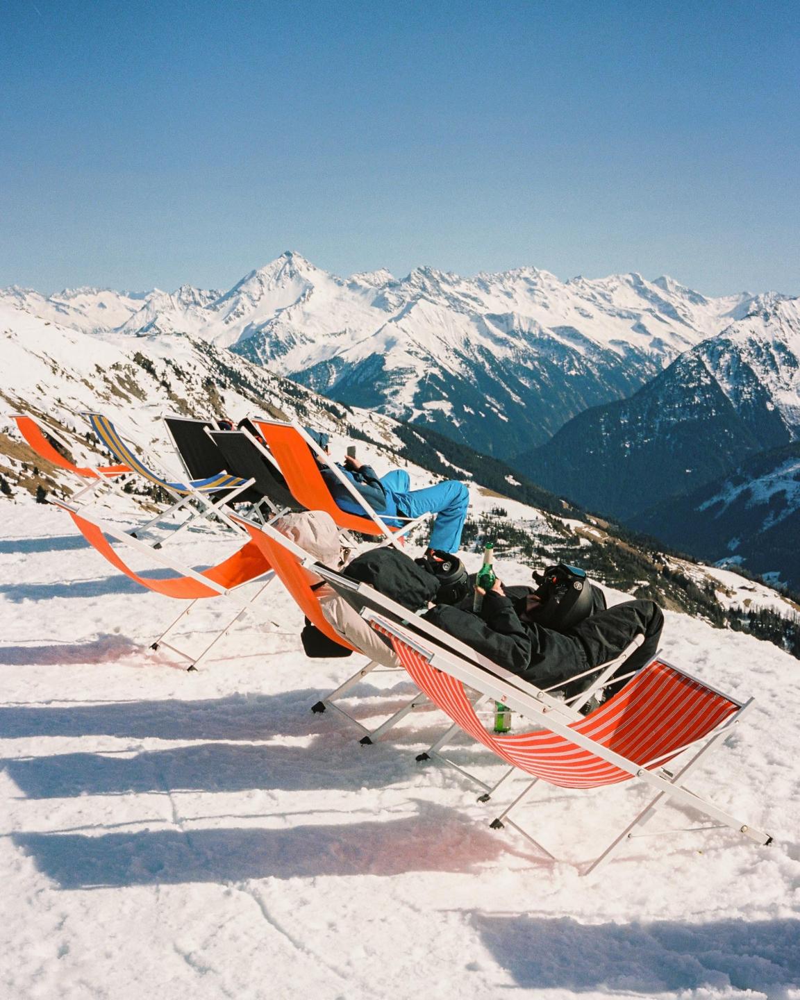 Several people relax in colorful deck chairs on a snowy mountain slope with a vast mountain range in the background.