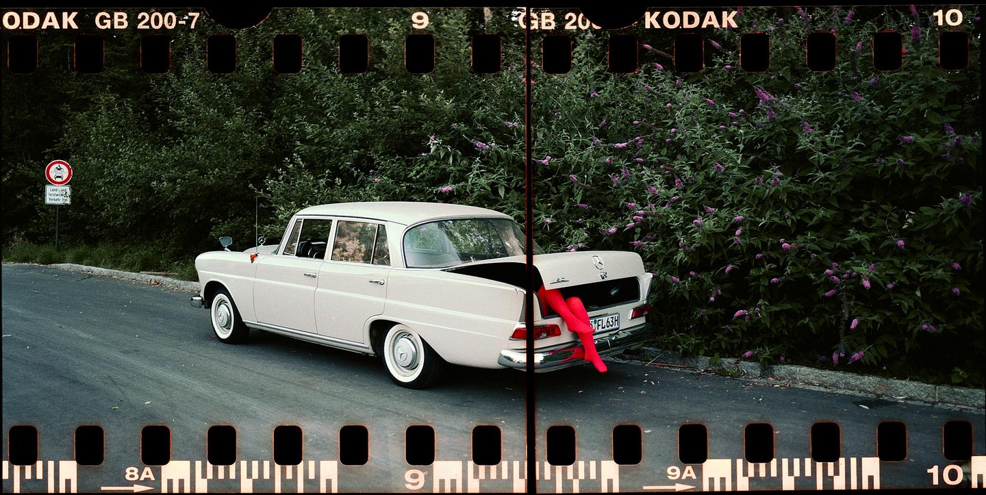 A white car with its trunk open and a pair of red legs sticking out is parked on the side of a road next to a bush.