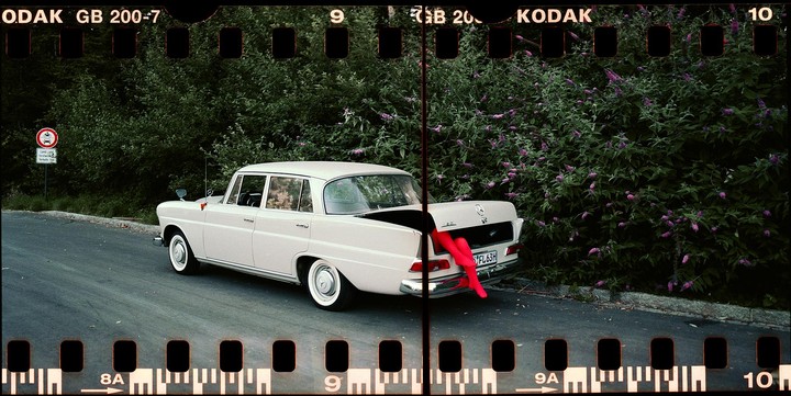 A white car with its trunk open and a pair of red legs sticking out is parked on the side of a road next to a bush.