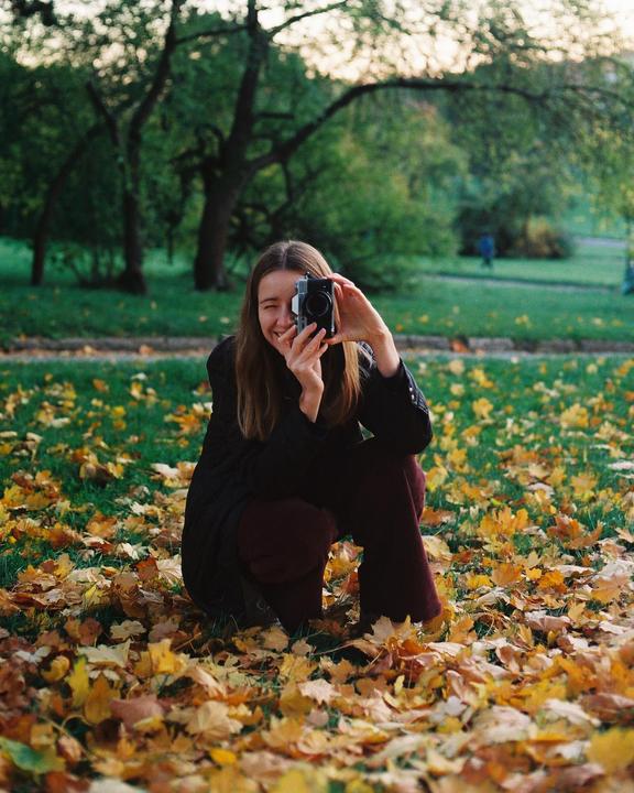 A woman crouches in a park covered in autumn leaves, holding a camera to her face and winking.