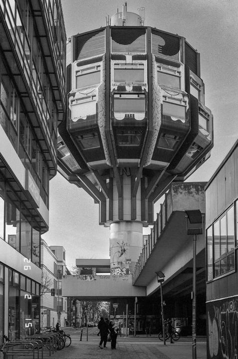 A unique hexagonal building on a single pillar stands between two other buildings in a city.