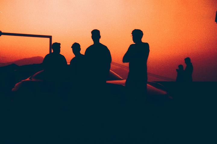 A group of men are silhouetted against a vibrant orange and red sky, with a car visible in the foreground.
