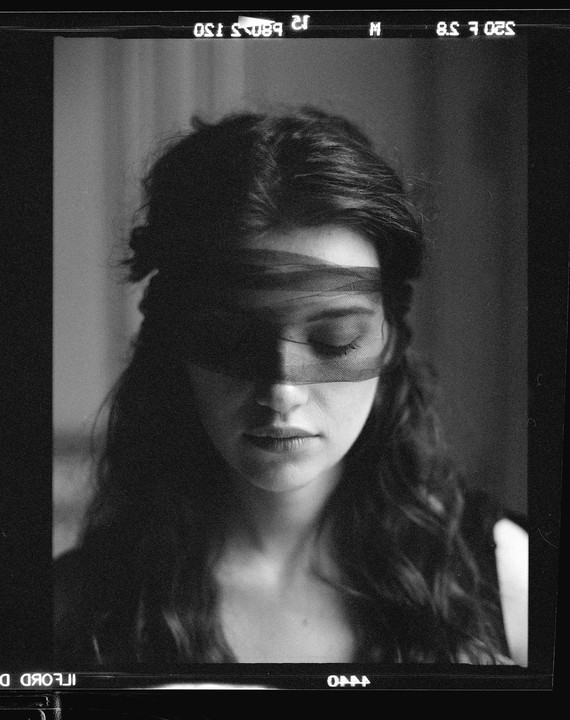A woman with long hair and a blindfold over her eyes looks down.
