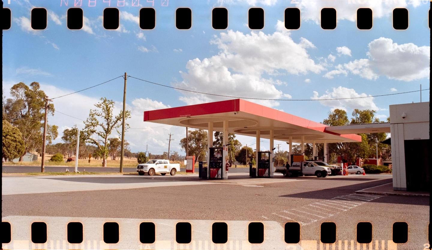 A gas station with a red canopy stands under a blue sky with white clouds, with several vehicles parked at the pumps.