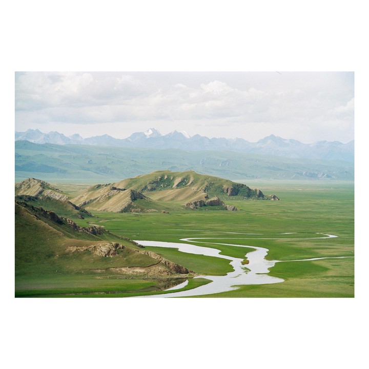 A winding river flows through a vast green valley with mountains in the distance under a cloudy sky.