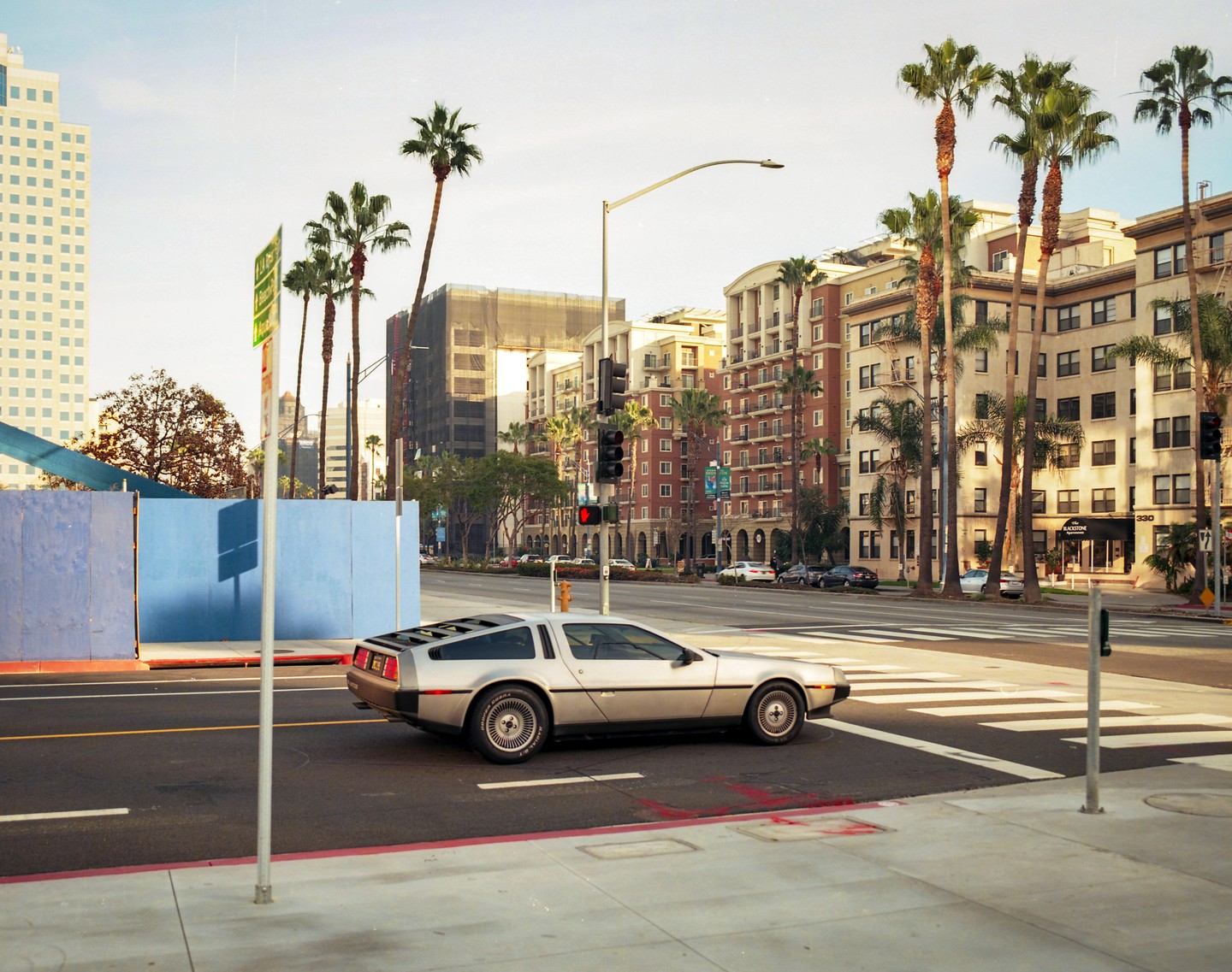 A silver DeLorean is stopped at a traffic light on a city street lined with palm trees and buildings.
