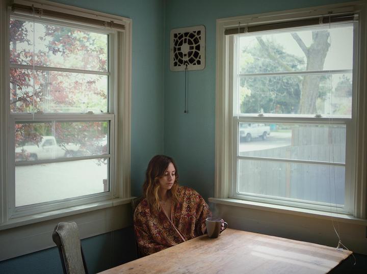 A woman in a patterned kimono sits at a wooden table with a steaming mug between two windows.