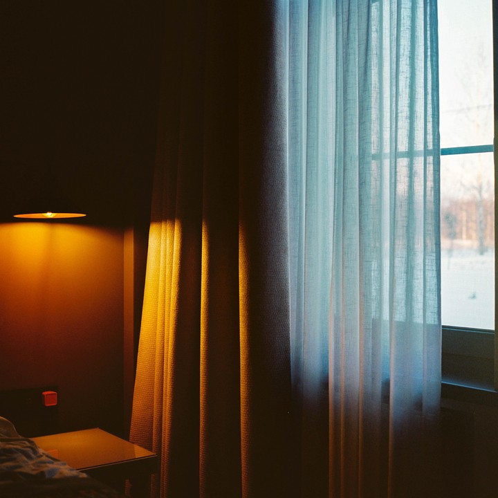 A hotel room with a window and a lamp on the wall is illuminated by natural light and artificial light.