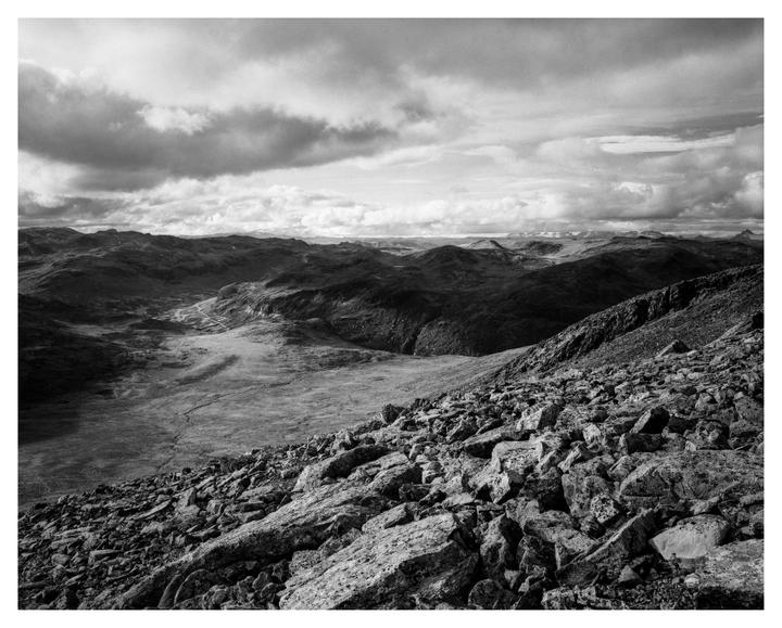 A black and white landscape shows a rocky slope in the foreground leading to a valley and distant mountains under a cloudy sky.