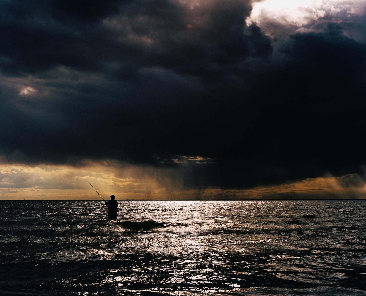 A man wades in the ocean with a fishing rod under a dramatic, cloudy sky.