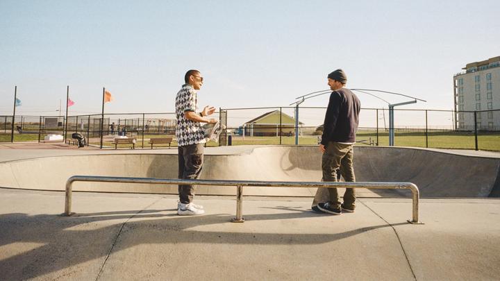 Two men stand in a skatepark, one holding a skateboard, with a building and flags visible in the background.