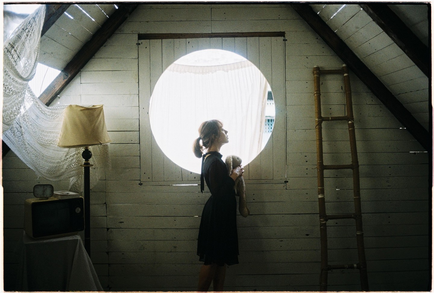 A girl holding a stuffed animal stands in front of a circular window in an attic room.