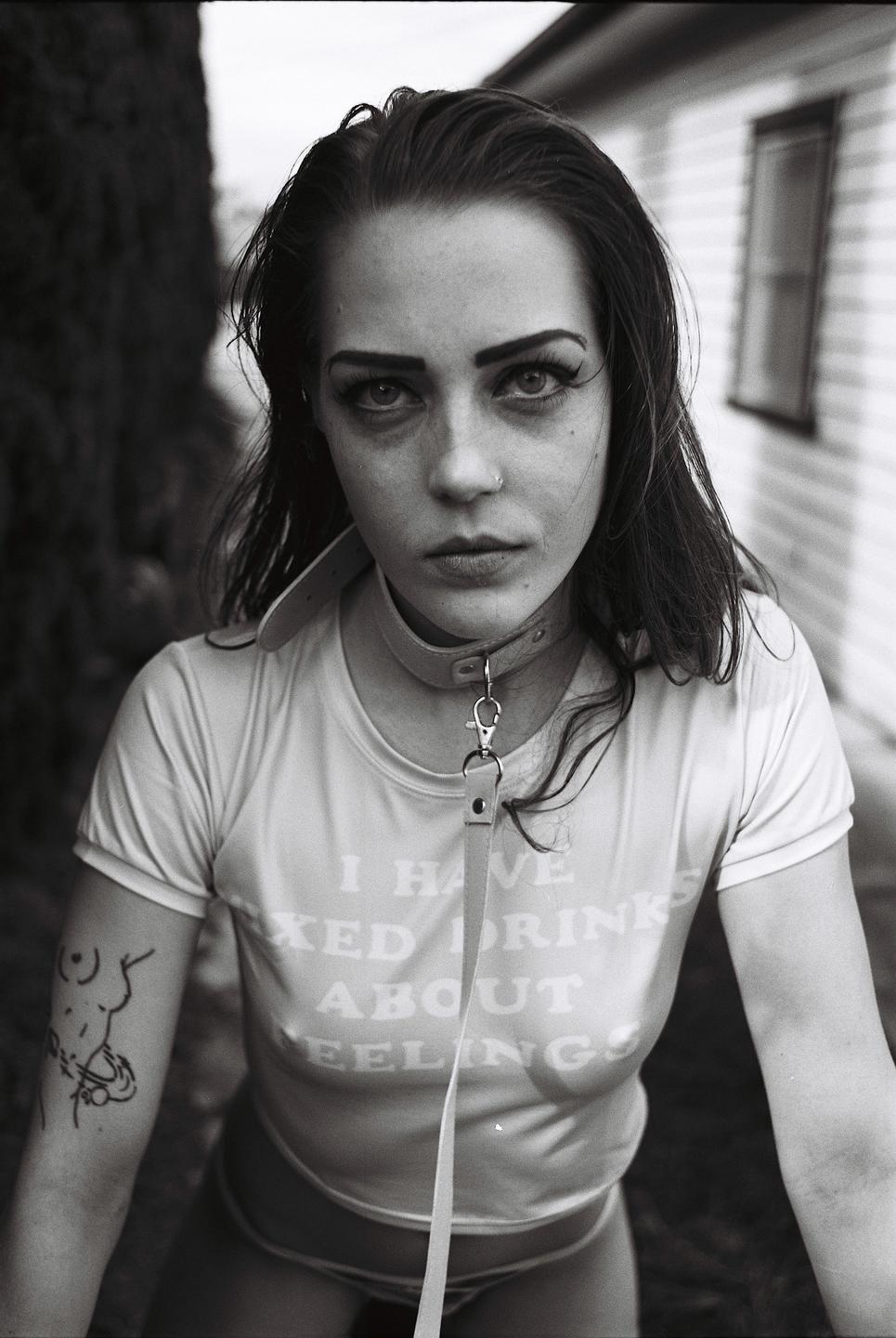 A woman wearing a collar and leash looks directly at the viewer.