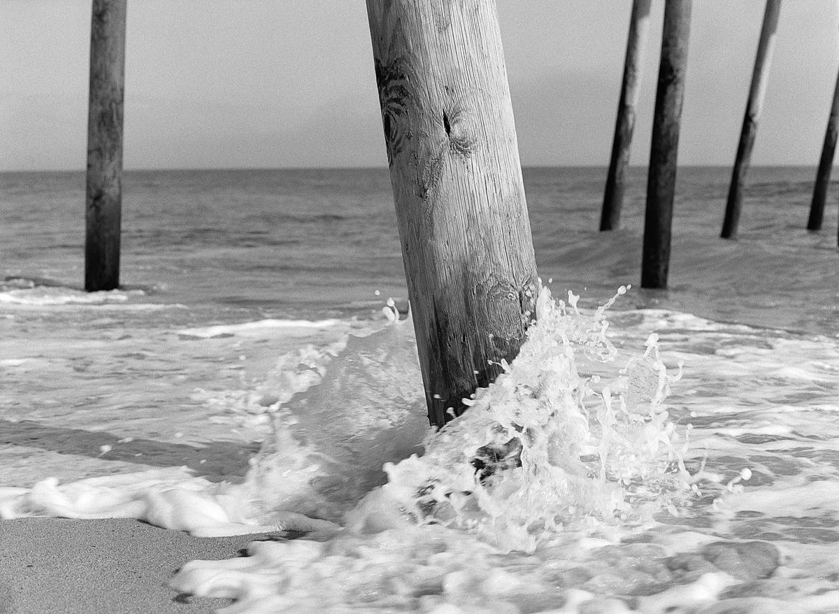 A wooden pier piling is splashed by a wave on a sandy beach.
