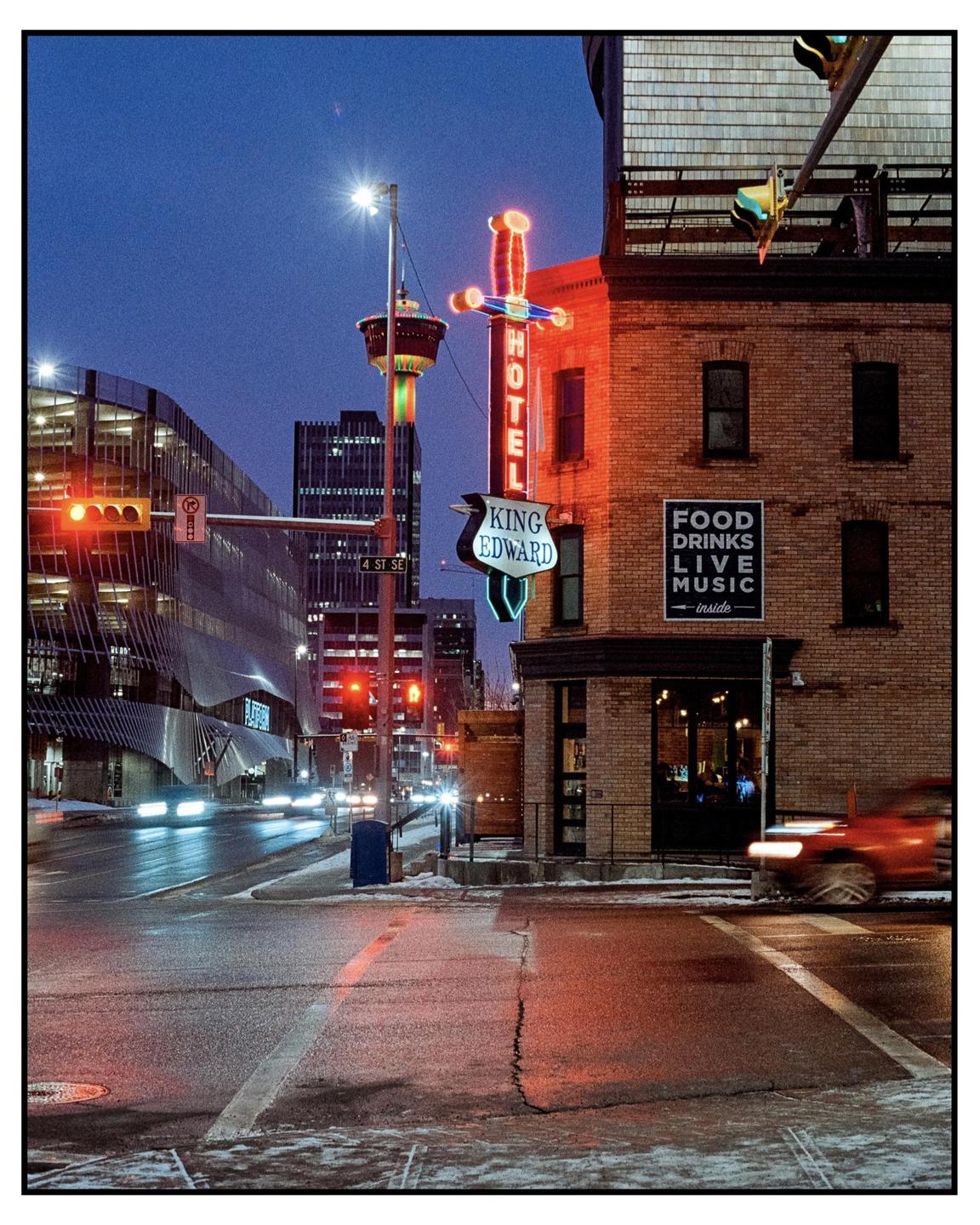 A neon sign for the King Edward Hotel glows brightly on a brick building at dusk, with car light trails on the street.