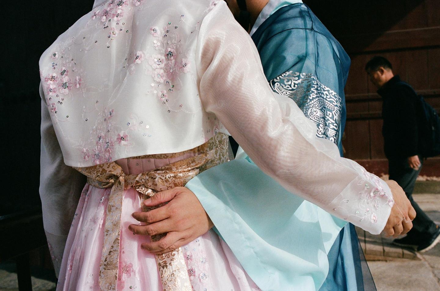 A couple wearing traditional Korean clothing stands close together, with a man walking in the background.
