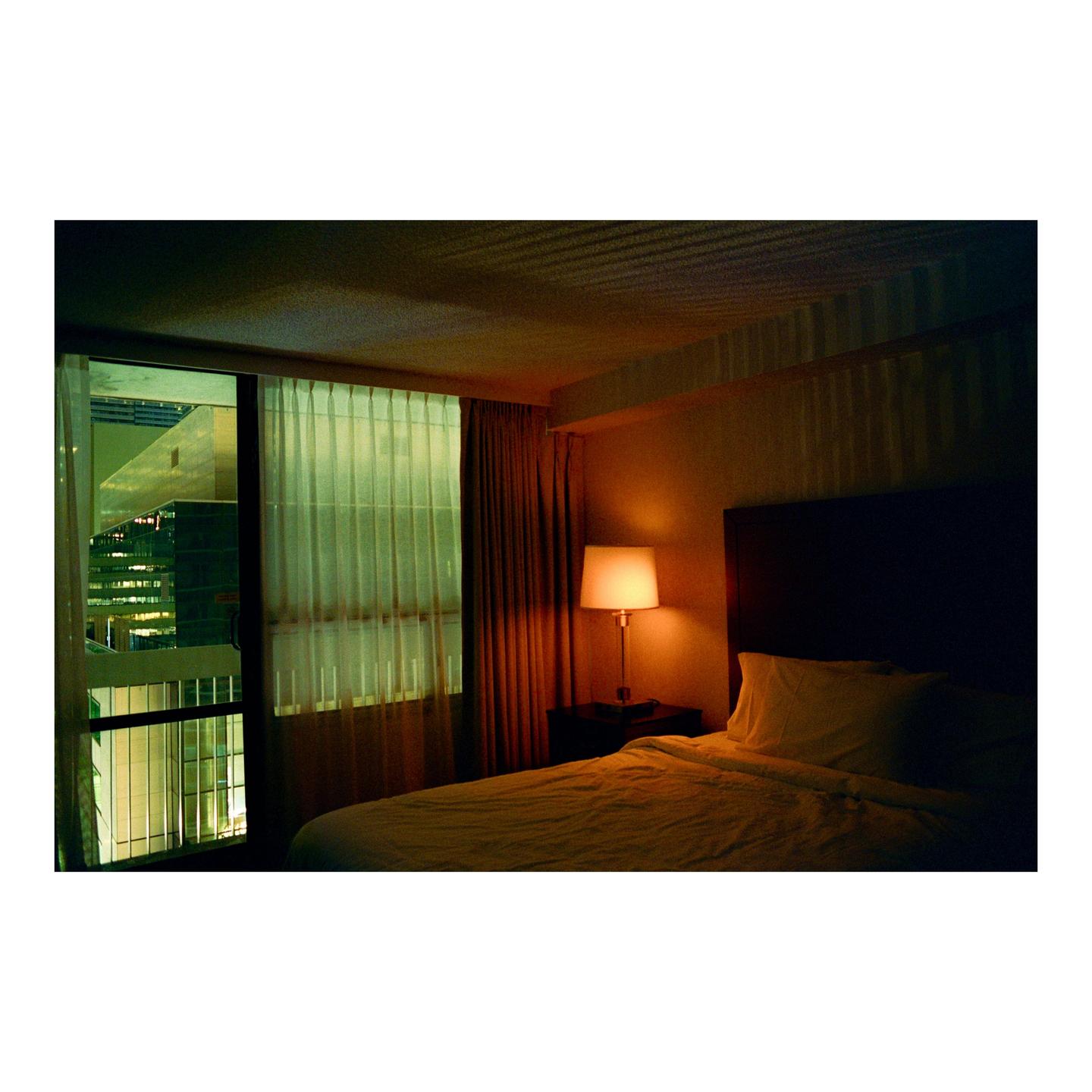 A hotel room at night features a bed, a lamp on a nightstand, and a window looking out onto city buildings.