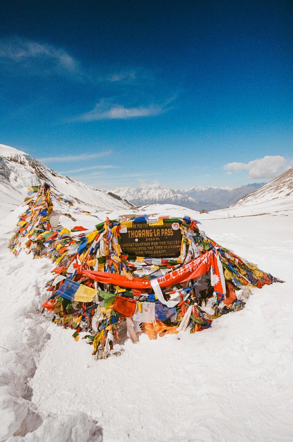A sign for Thorung La Pass is surrounded by colorful prayer flags in a snowy mountain landscape under a bright blue sky.