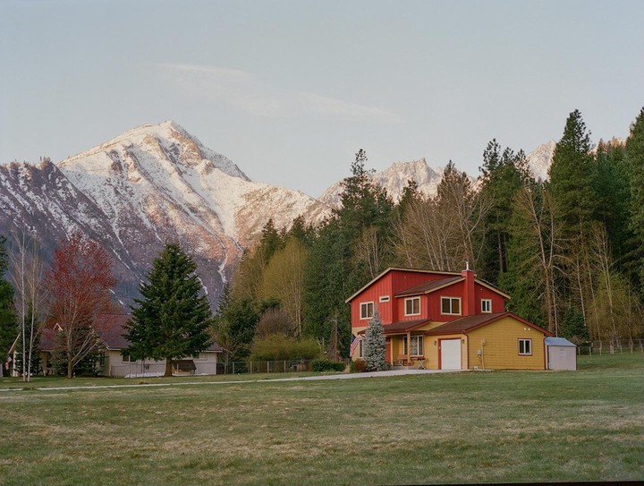 A red and yellow house sits in a grassy field with a snow-capped mountain and forest in the background.