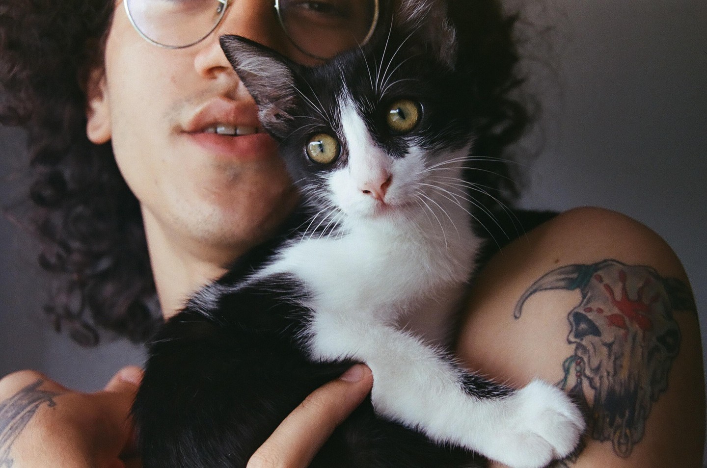 A man with curly hair and glasses holds a black and white kitten with green eyes, revealing a skull tattoo on his shoulder.