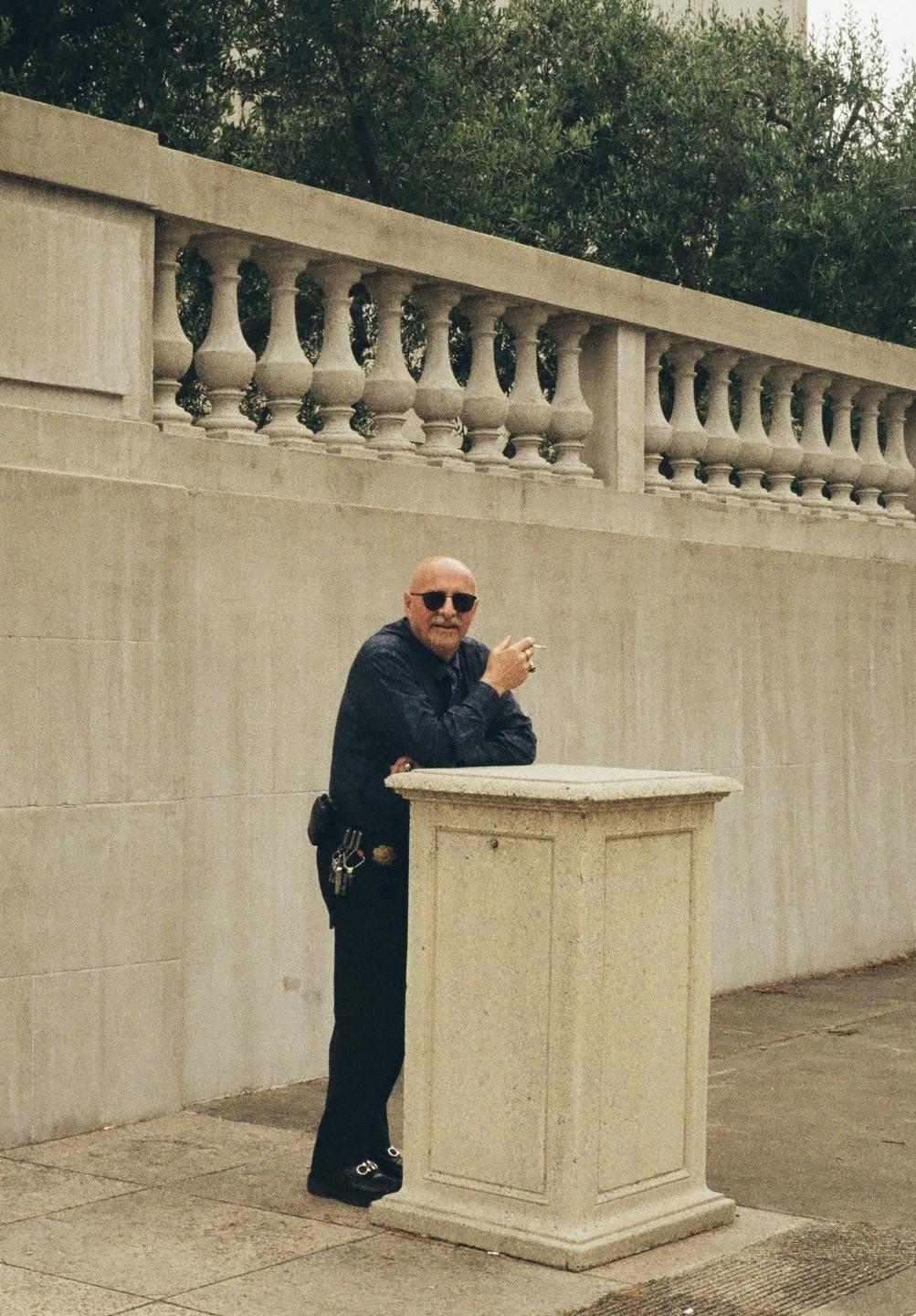 A man with a beard and sunglasses leans on a stone pedestal while holding a cigarette.