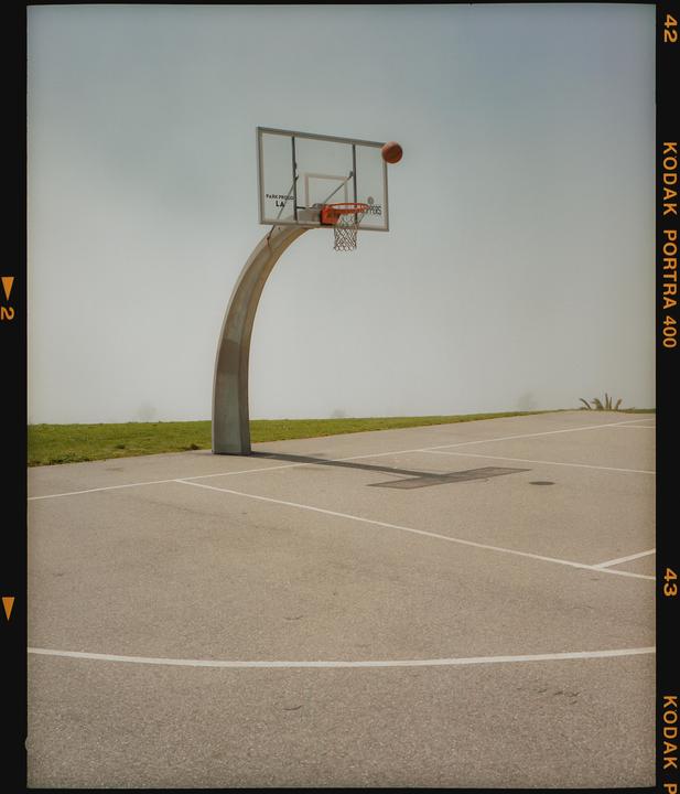 A basketball is in mid-air above a basketball hoop on an outdoor court.
